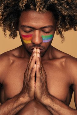 Calm shirtless black man with rainbow on cheeks and curly hair clasping hands and praying with closed eyes against brown background
