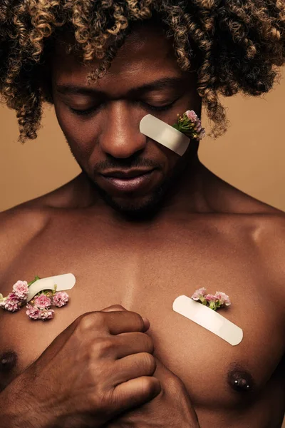 Vulnerable shirtless African American male with fresh patches attached to damaged skin with patches clasping hands on chest against brown background