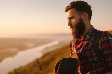 Bearded man in checkered shirt with hat looking away and admiring river at sundown in countryside