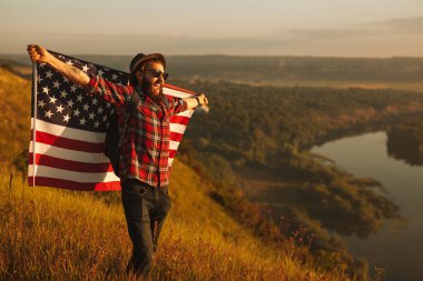 Cheerful bearded man in checkered shirt with hat and sunglasses, spreading USA flag behind back and screaming while standing on grassy hill near river at sunset
