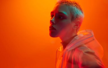 Young androgynous woman in futuristic raincoat with short hair looking away while standing under bright neon light against orange background