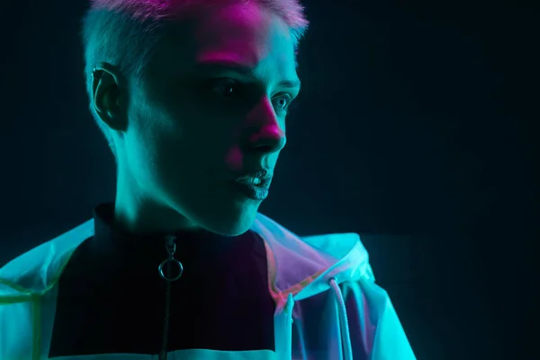 Crop cyberpunk woman with short blond hair in hooded raincoat looking away thoughtfully against black background in dark studio with neon lights
