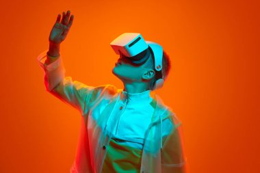 Concentrated young female with short hair in transparent raincoat looking at raised arm while experiencing virtual reality in modern headset against orange background