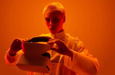 Young woman in futuristic raincoat putting on VR glasses and looking at camera under bright orange neon light