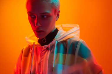 Young female model in translucent futuristic coat with short hair looking away under neon illumination against orange background