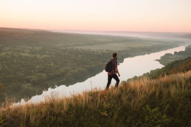 Man in checkered shirt with backpack walking on hill slope near calm river while travelling in countryside alone at sunset