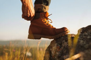 Unrecognizable man stepping on rough stone and adjusting leather boot during trip through field on autumn day