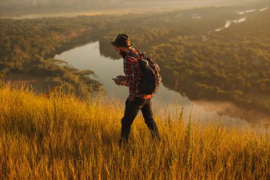 High angle of bearded man in checkered shirt with hat and backpack, strolling on grass near calm river and browsing cellphone in morning in countryside