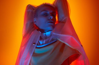 Young woman in futuristic clothes with short hair raising arms and looking at camera under colorful neon illumination