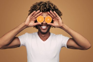 Happy African American man in white t shirt smiling and holding haves of fresh orange near eyes while being on diet against brown background