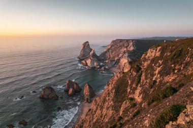 Drone view of rocky cliffs surrounding Ursa Beach near waving sea against sundown sky in Portugal