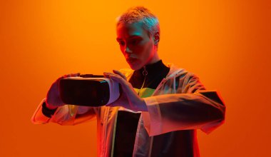 Young woman in futuristic outfit and short hair inspecting VR glasses before playing videogame under orange neon illumination