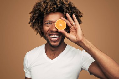 Beyaz kıvırcık saçlı mutlu Afro-Amerikan erkek kameraya gülümsüyor ve taze portakalın yarısını gözüne yakın tutuyor. Sağlıklı yaşam tarzını kahverengi arka plana karşı teşvik ediyor.