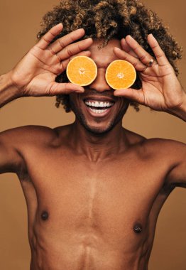 İyimser, üstsüz, Afro-Amerikan erkek. Elinde doğal turuncu gözleri var. Kahverengi arka planda eğlenirken gülümsüyor.