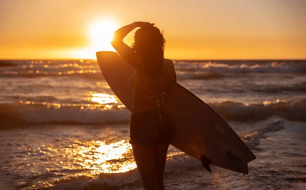 Anonymous woman with surfboard touching head and admiring bright sunset sky while standing on beach near waving sea
