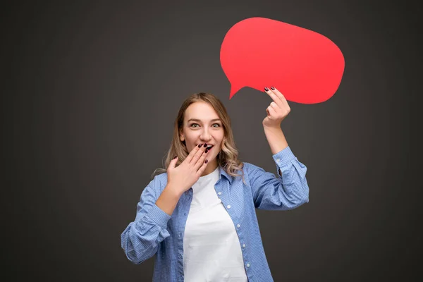 Amazed young female in casual clothes showing blank red speech bubble and covering mouth while sharing secret against black background