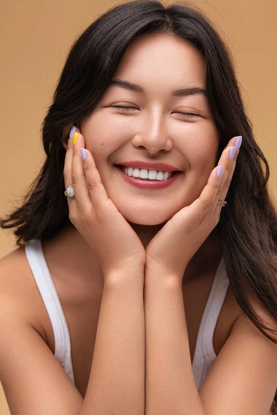 Cheerful young Asian female model with dark hair smiling with closed eyes and touching cheeks during skin care routine against beige background