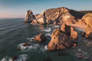Praia da Ursa 'da bulutsuz gün batımına karşı köpüklü okyanus dalgalarıyla yıkanan devasa kayalık uçurumların nefes kesici manzarası.