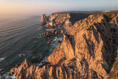 Portekiz 'de gün batımına karşı dalgalı denizin kıyısındaki dalgalı kayaların ve Ursa sahilinin resimli hava manzarası