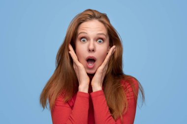 Kızıl saçlı, uzun saçlı, yanaklara dokunan ve ağzı açık bir şekilde kameraya bakarken mavi arka planda şaşırmış bir kadın.