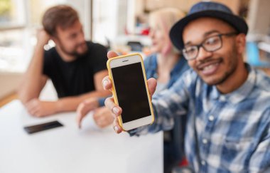 Afro-Amerikan erkeğin gündelik kıyafetleriyle kafeterya masasında arkadaşlarının yanında otururken siyah ekranlı cep telefonu göstermesi ve reklam uygulaması.
