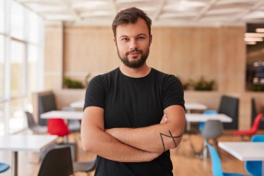 Siyah tişörtlü, kendine güvenen bir erkek çalışan kollarını kavuşturup çalışma alanının bulanık arka planında kameraya bakıyor.