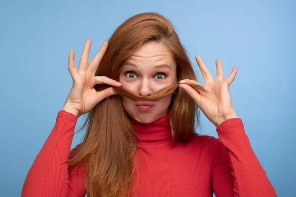 Funny redhead female in red turtleneck pouting lips and looking at camera while making mustache from long hair against blue background