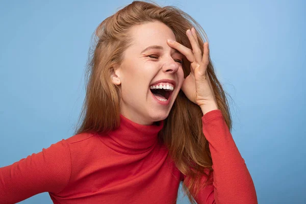 Happy young female in red turtleneck with ginger hair touching forehead and laughing with closed eyes against blue background