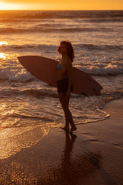 Full body woman with surfboard standing on wet sand near waving sea at sundown during summer vacation on beach