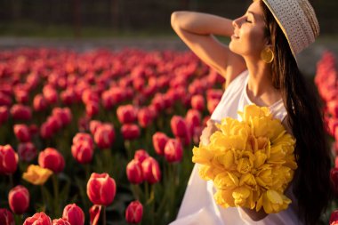 Beyaz elbiseli, sarı lale desenli hasır şapkalı, koyu renk saçlı ve kırsal kesimde bahar sabahı kapalı gözlerle güneş ışığının tadını çıkaran pozitif İspanyol bir kadın.