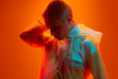 Şeffaf fütüristik yağmurluk giymiş genç bir kadın model turuncu arka planda renkli neon ışığı altında dururken kısa saçlara dokunuyor.