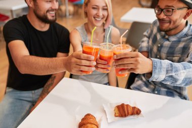 Restoranda kahvaltıda kruvasanlarla masada otururken gülümseyen ve plastik bardakta taze havuç suyu tokuşturan çok kültürlü kadın ve erkeklerden.