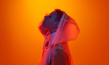 Kısa saçlı, kafaya dokunan ve şeffaf fütüristik yağmurluğun kapüşonunu çıkartan kadın modelin yan görünüşü, turuncu arka plana karşı renkli neon aydınlatma altında dururken,