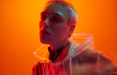 Parlak neon ışığın altında turuncu arka planda dururken yarı saydam yağmurluklu, kısa saçlı fotomodelin teki.