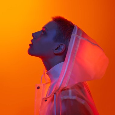 Kapüşonlu yağmurluk giyen, parlak neon ışığı altında turuncu arka planda görünen fütürist genç kadın modelinin profili.