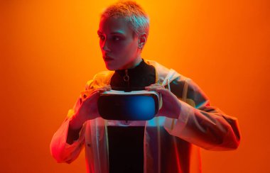 Fütüristik kıyafetli ciddi bir kadın stüdyoda turuncu arka plana bakarken çağdaş sanal gerçeklik gözlükleriyle ayakta duruyor.