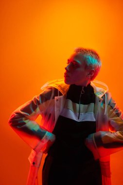 Ciddi bir kadın, kısa sarı saçlı, siber punk tarzı giyinmiş. Stüdyoda elleri belinde turuncu arka planda dikiliyor ve gözlerini kaçırıyor.