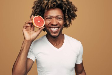 Kıvırcık saçlı neşeli Afro-Amerikan erkek, kameraya gülümseyerek bakıyor ve C vitamini dolu greyfurtun yarısını gösterirken kahverengi arka planda sağlıklı yaşam tarzını teşvik ediyor.