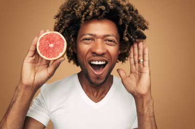 Kıvırcık saçlı, kollarını kaldıran ve kameraya bakan beyaz tişörtlü neşeli Afro-Amerikan erkek ağzı açık bir şekilde kahverengi arka planda taze greyfurtun yarısını gösteriyor.