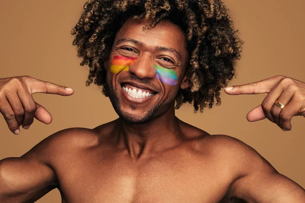 Happy black curly man pointing at multicolored rainbow flags painted on cheeks while smiling at camera on brown background