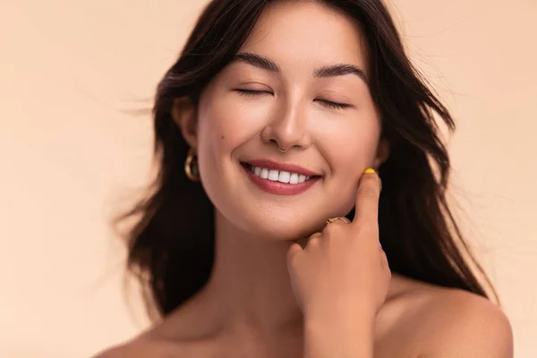 Cheerful Asian female model with smooth skin and brown hair touching face for concept of beauty routine on beige background