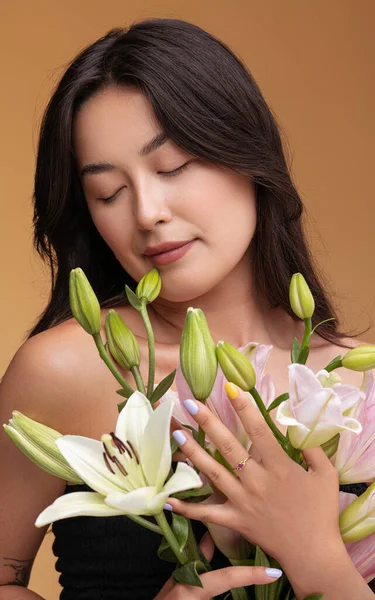 Tranquil young Asian female model with long dark hair and closed eyes standing against beige with bunch of fresh gentle white lilies in hands