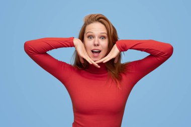 Balıkçı yaka yakalı kızıl saçlı güzel bir manken kameraya bakıyor ve stüdyoda mavi arka planda yüzlere dokunuyor.