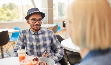 Kareli gömlekli, şapkalı, gülümseyen ve kız arkadaşını dinleyen mutlu bir Afro-Amerikan erkeği. Gündüz vakti güneşli bir restoranda, masada otururken.