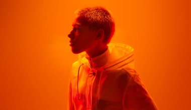 Kısa saçlı, siber punk kıyafetli, turuncu arka planda neon ışığıyla stüdyoda duran duygusuz bir kadının yan görüntüsü.