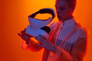 Sanal gerçekliğin geleceksel gözlüğünü kullanarak turuncu arka planda, stüdyoda neon ışıklı sanal gerçekliği keşfeden ciddi bir kadın.