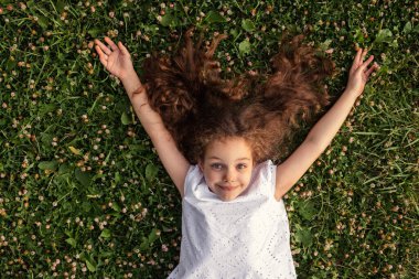 Kahverengi saçlı, neşeli çocuk gülümseyen ve kollarını kaldıran kameraya bakarken yaz günü parkta yonca çiçekleriyle yeşil çimlerin üzerinde uzanırken.