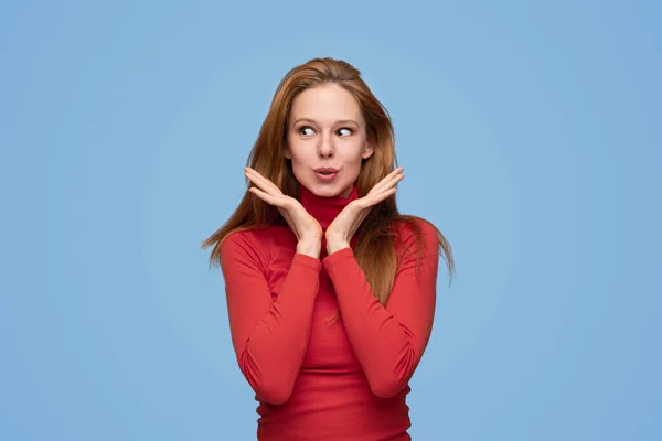 Young content female in red turtleneck pouting lips and standing with hands near face on blue background in studio