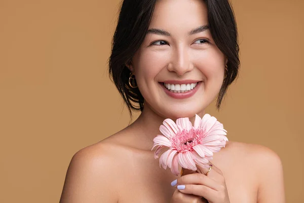 幸せな若いですアジアの女性とともに裸肩保持新鮮なピンクのガーベラの花と離れて見ますおもちゃの笑顔で茶色の背景 — ストック写真