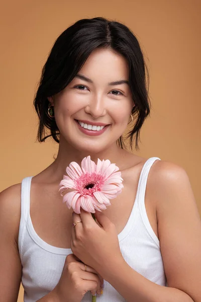 ポジティブな若いですアジアの女性で白い上に自然ピンクのガーベラの花見てカメラで笑顔で茶色の背景 — ストック写真